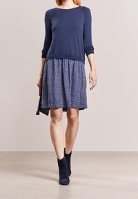 Haut en tricot marine à manches longues et jupe plissée arborant un motif géométrique bleu. Associé à des bottines marine et tenant une pochette.