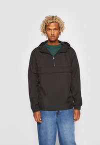 Sort, letvægts pulloverjakke med hætte, halv-lynlås foran og elastiske manchetter, båret over en grøn t-shirt og blå jeans.