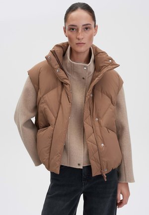Brown puffer vest with a high collar, quilted design, and snap-button closure, layered over a beige top and paired with dark jeans.