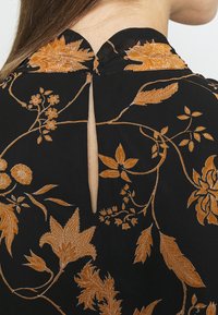 Vêtement noir avec broderie florale orange, doté d'un col rond et d'une ouverture en forme de clé à l'arrière. Texture de tissu douce évidente.