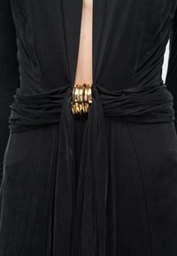 Schwarzer geraffter Stoffgürtel mit übereinanderliegenden glänzenden goldenen Metallringen, die an der Taille über einem schwarzen Kleidungsstück mit offenem Vorderteil befestigt sind.