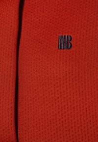 Röd strukturerad tyg med ett subtilt mönster. Har en liten broderad logotyp i marinblått. Slät yta med ett luftigt utseende.