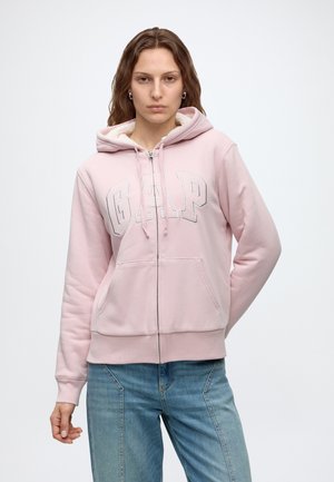 Frau trägt eine hellrosa GAP Kapuzenjacke mit Reißverschluss und Fronttaschen sowie hellblaue Jeans, steht mit einer Hand hinter dem Rücken.