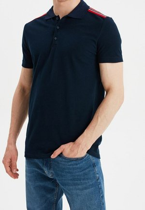Mand iført navyblå polo med røde og hvide striber på skuldrene og blå jeans, den ene hånd i lommen.