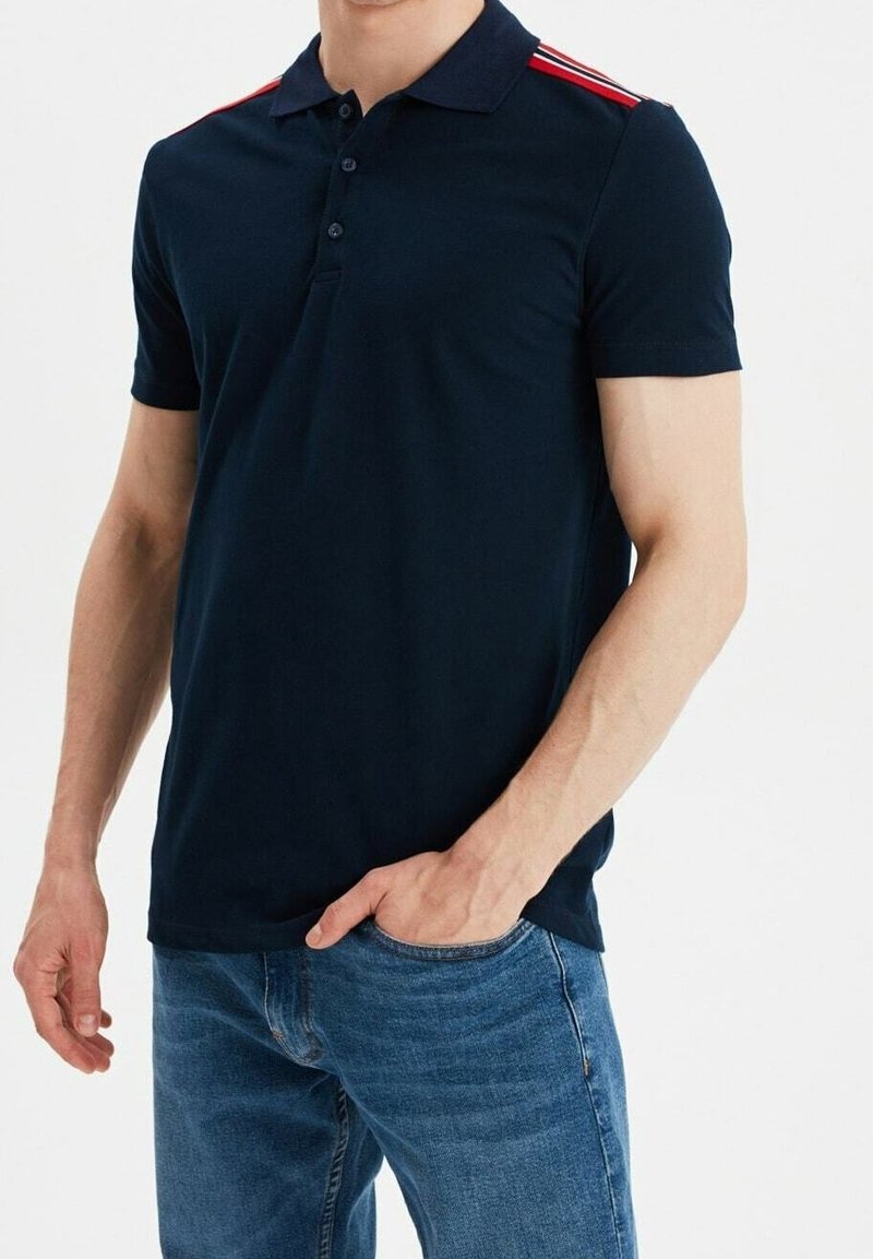 Homme portant un polo bleu marine avec des rayures rouges et blanches sur les épaules et un jean bleu, une main dans la poche.