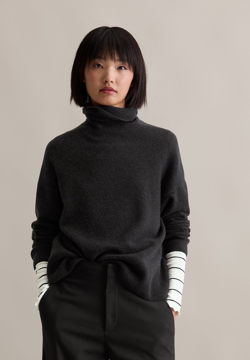 Schwarzer Rollkragenpullover aus weichem Material mit lockerer Passform, kombiniert mit einem weißen Langarmunterhemd mit schwarzen Streifen.