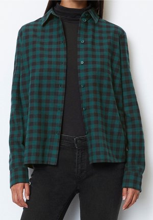 Kobieta ubrana w ciemnozieloną i czarną kratowaną koszulę zapinaną na guziki, nałożoną na czarny golf i czarne dżinsy, stojąca na białym tle.