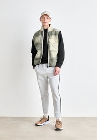 Γιλέκα φλις με καρό μοτίβο σε πράσινο και μπεζ, μαύρη μπλούζα, ανοιχτό γκρι παντελόνια φόρμας με πλάγιες λωρίδες και καφέ αθλητικά παπούτσια.