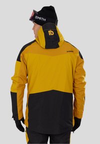 Skijas in geel en zwart, met een grote capuchon, verstelbare manchetten en zakken. Gemaakt van waterdicht materiaal met een gladde textuur.