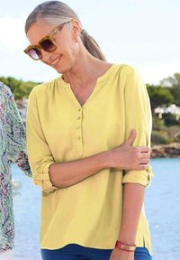 Blusa gialla chiara con scollatura a V e patta di bottoni. Maniche lunghe arrotolate, texture morbida e leggero lucido. Indossata con pantaloni blu.