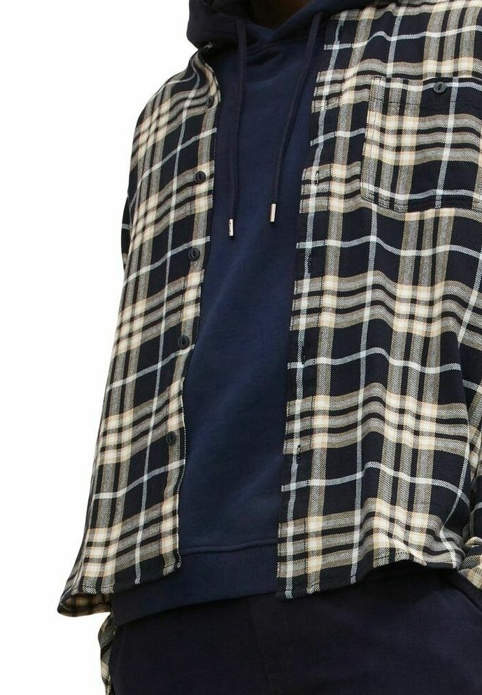 Homme portant un sweat à capuche bleu marine sous une chemise à carreaux noire, blanche et beige, déboutonnée, à manches longues.