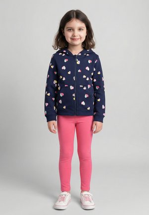 Junges Mädchen steht, trägt einen marineblauen Kapuzenpullover mit Reißverschluss und bunten Herzmustern, pinke Leggings und weiße Turnschuhe.