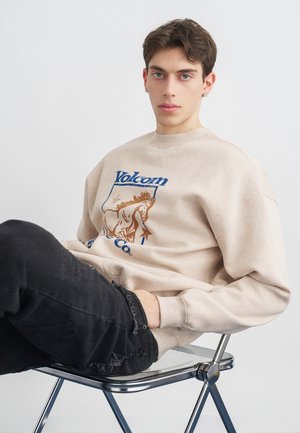 Jeune homme aux cheveux foncés portant un sweat-shirt beige Volcom et un jean noir déchiré, assis sur une chaise transparente devant un fond gris uni.