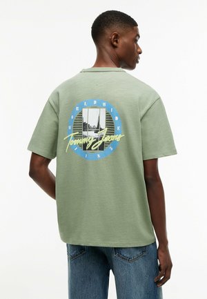 Homme portant une chemise manches courtes vert sauge avec un texte graphique bleu et jaune « Tommy Jeans » et une image dans le dos, associée à un jean bleu.