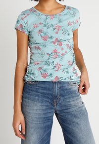 Top de manga corta de color azul claro con patrones florales en rosa y verde. Tela suave, diseño ajustado, cuello redondo, combinado con jeans de azul claro.