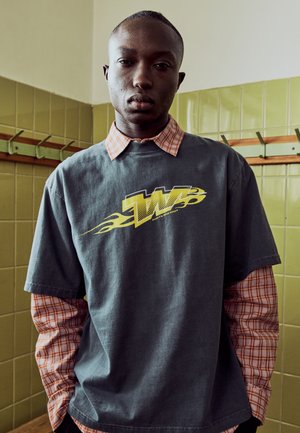 Ung mand står i et flisebelagt rum iført en grå T-shirt med et gult motiv over en rød ternet skjorte med hænderne i lommerne.