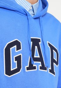 Približen posnetek osebe, ki nosi modro kapuco GAP preko modre in bele srajce s črtami in gumbi.