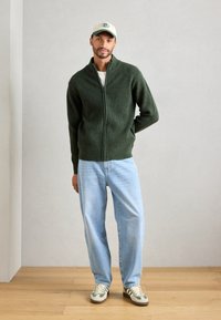 Mann iført en grønn zip-up genser, lyseblå jeans, joggesko og en beige caps, stående på tregulv mot en enkel lys vegg.