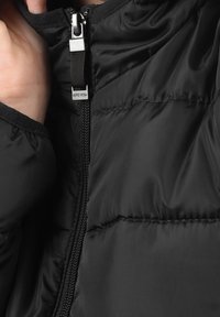 Schwarze Daunenjacke mit einem Reißverschluss vorne, gesteppter Textur und weichem Material. Etikett in der Nähe des Reißverschlusses sichtbar. Schlanke Passform mit hohem Kragen.