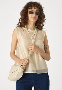 Beige sleeveless blouse with a sheer texture, round neckline, paired with a light-colored handbag and pearl necklace, dark denim pants.