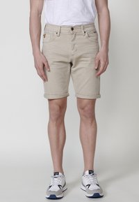 Man med beige upprullade shorts, vit t-shirt och vita sneakers som står mot en enkel ljus bakgrund.