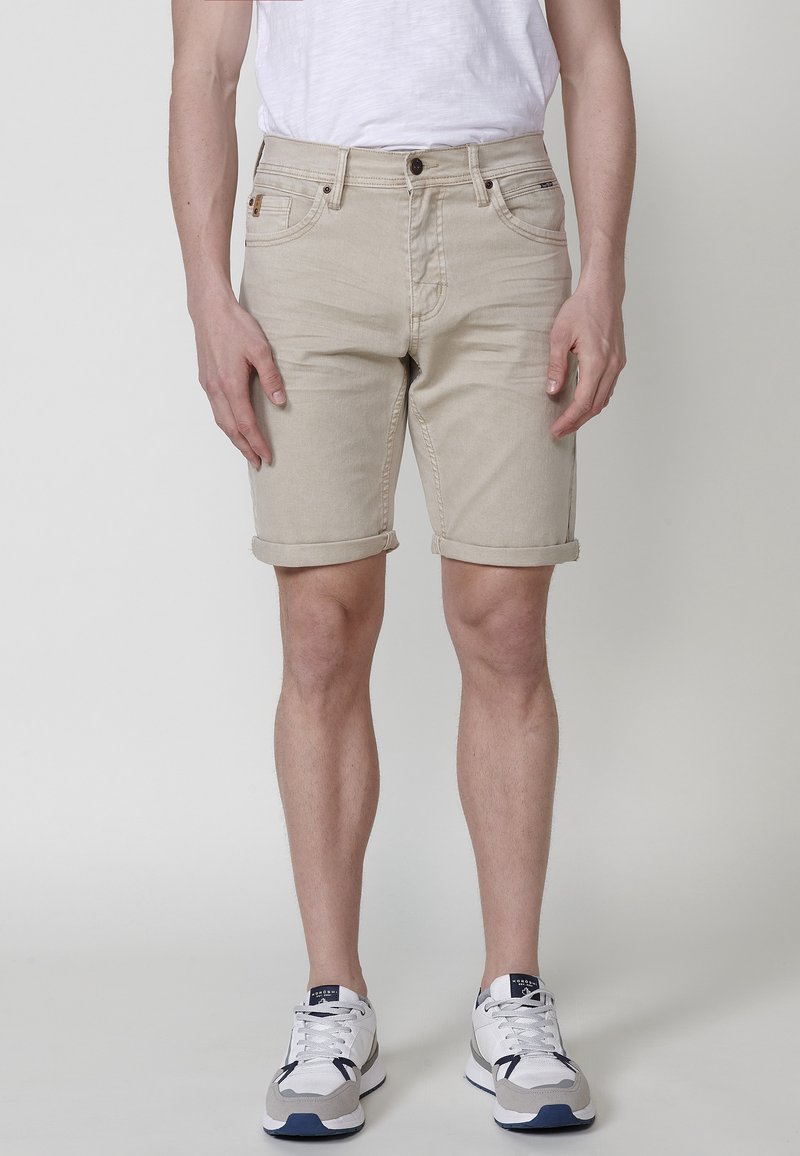 Man med beige upprullade shorts, vit t-shirt och vita sneakers som står mot en enkel ljus bakgrund.
