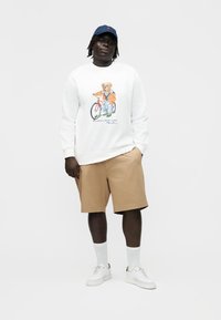 Mann trägt ein weißes Sweatshirt mit einem Bären auf dem Fahrrad als Grafik, beige Shorts, weiße Turnschuhe, weiße Socken und eine blaue Kappe, steht vor weißem Hintergrund.