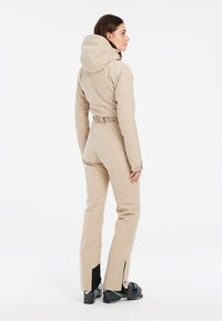 Completo da sci beige con vita aderente, cappuccio e aperture con zip alle caviglie. Presenta un tessuto liscio e dettagli neri ai polsini e agli stivali.