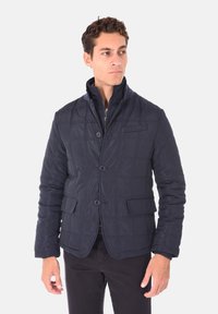Giacca trapuntata navy con colletto alto, due tasche frontali e bottoni. Realizzata in tessuto leggero e liscio con una finitura leggermente lucida.