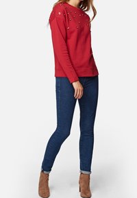 Roter Sweatshirt mit dekorativen Verzierungen an den Schultern, kombiniert mit blauen Skinny Jeans und beigen Stiefeletten, was einen lässigen Stil zeigt.