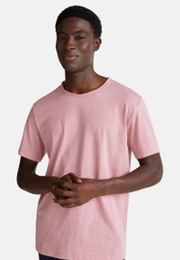 Joven hombre negro con una camiseta de manga corta de color rosa claro, de pie con las manos entrelazadas a la altura de la cintura, frente a un fondo blanco liso.