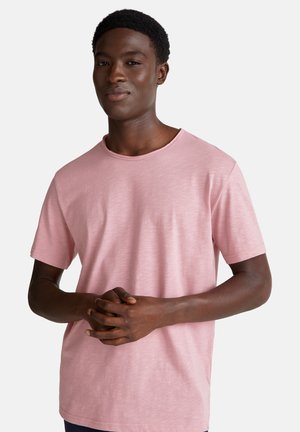 Junger schwarzer Mann in einem hellrosa kurzärmeligen T-Shirt, steht mit gefalteten Händen auf der Taille vor einem schlichten weißen Hintergrund.