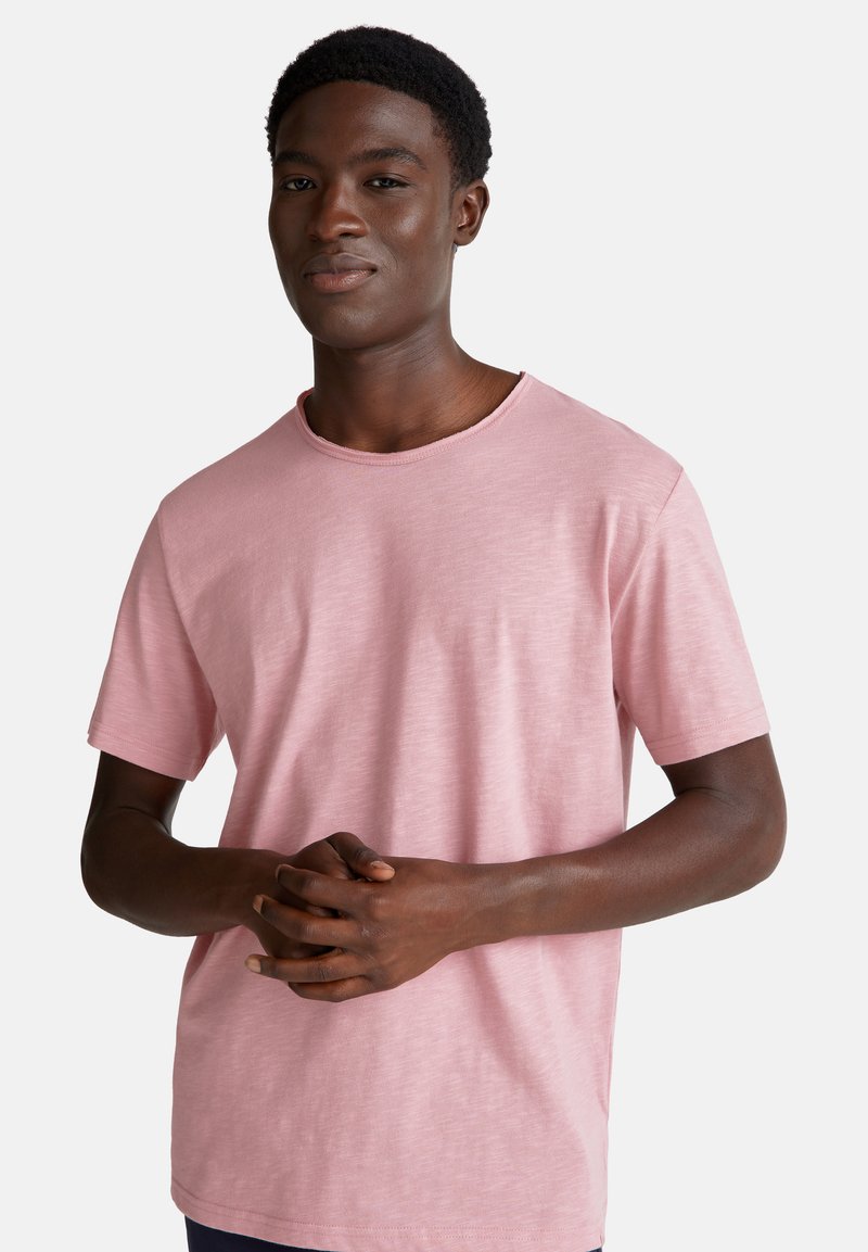 Joven hombre negro con una camiseta de manga corta de color rosa claro, de pie con las manos entrelazadas a la altura de la cintura, frente a un fondo blanco liso.