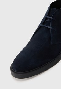 Cizme chukka din suede bleumarin, cu vârful rotund, talpă din cauciuc negru și șireturi închise, pe un fundal alb.
