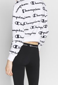 Osoba ubrana w biały krótki sweatshirt i czarne legginsy, oba z powtarzającym się nadrukiem logo Champion, widziana od tyłu.