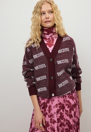 Blond vrouw die een bordeauxrode cardigan met witte tekst draagt over een roze bloemencoltrui en bijpassende geplooide rok, staand tegen een effen achtergrond.