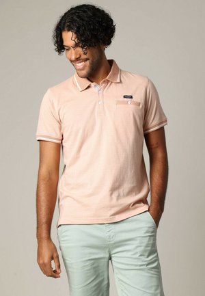 lichtroze polo shirt van zacht stof, met een kraag, drie knopen en een borstzak met een label. Gecombineerd met lichtgroene broek.