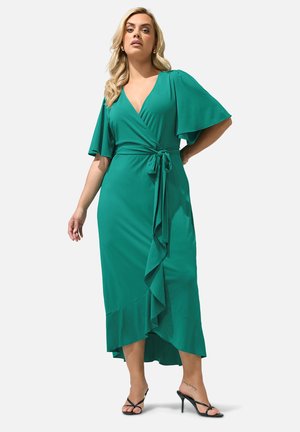 Femme portant une robe portefeuille verte avec des manches flottantes et un ourlet à volants, associée à des sandales à talons noirs, debout avec confiance.