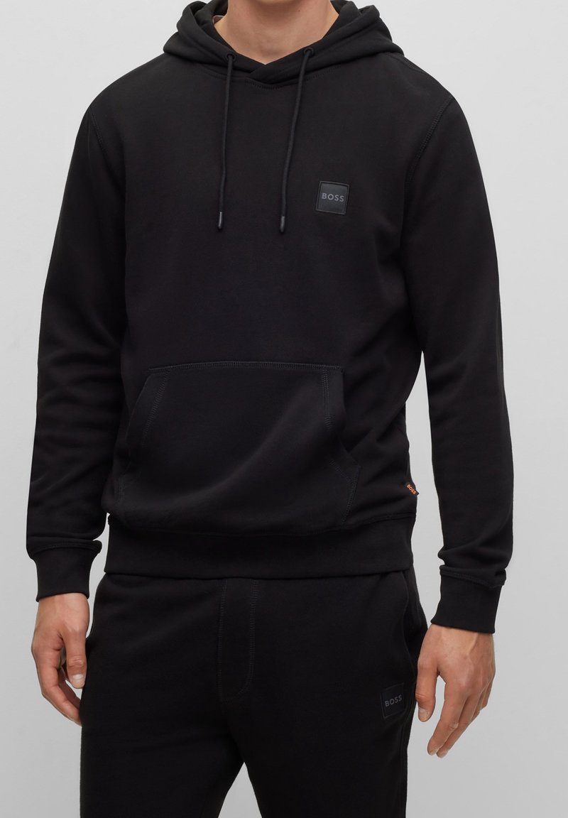 Svart hoodie i mjukt material med en framficka, dragsko på huvan och en liten märkeslapp på bröstet. Slät textur, avslappnad passform.