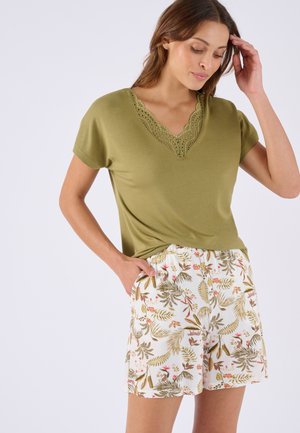 Vrouw draagt een olijfgroene V-hals top met kantdetails en witte shorts met een meerkleurig bloem- en bladerenpatroon, hand in de zak.