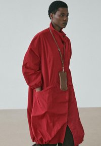 Roter, übergroßer Mantel mit gepufften Ärmeln und elastischen Bündchen. Verfügt über eine Knopfleiste vorne und zwei Seitentaschen. Inklusive einer braunen Zubehörtasche.