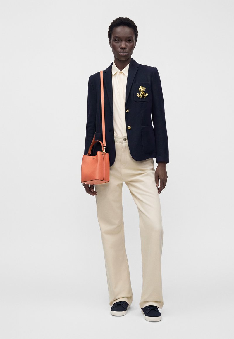 Γυναίκα που φοράει μπλε σκούρο σακάκι με χρυσό έμβλημα, κρεμ πουκάμισο και παντελόνι, μαύρα αθλητικά παπούτσια, κρατώντας κοραλλί τσάντα ώμου.