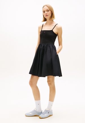 Frau steht in einem schwarzen ärmellosen Kleid mit Taschen, weißen Socken und hellblauen Turnschuhen vor einem schlichten weißen Hintergrund.