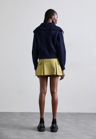 Navy sweater with a ribbed collar, paired with a pleated olive-green mini skirt. Black chunky shoes complete the outfit.