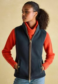 Navy modrá fleece vesta s vysokým golierom, vybavená zlatým zipsom a dvoma bočnými vreckami so zlatými akcentmi, nosená cez rebrovaný oranžový sveter.