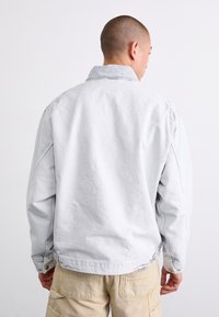 Lys grå denimjakke med en afslappet pasform, knapmanchetter og en foldet krave. Har synlige sømme og en subtil tekstur.