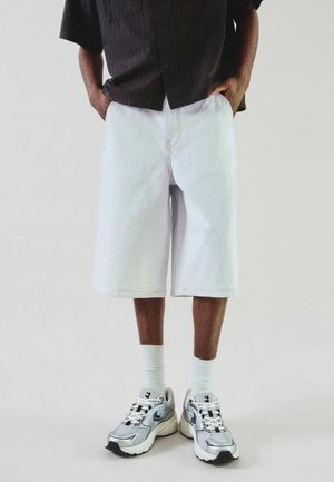 Személy világos farmer térd hosszúságú rövidnadrágot, fehér bokazoknit, ezüst-fehér sportcipőt és sötét rövid ujjú, gombos inget visel.