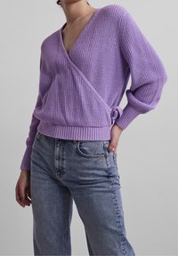 Fioletowy sweter z dzianiny o luźnym kroju i głębokim dekolcie w kształcie litery V, z prążkowanymi mankietami i wiązaniem z boku, w zestawieniu z jasnoniebieskimi dżinsami.