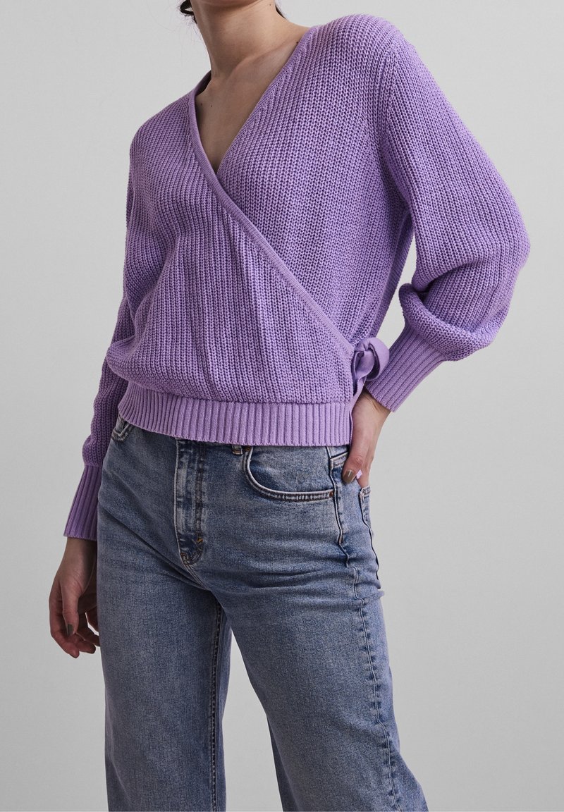 Fioletowy sweter z dzianiny o luźnym kroju i głębokim dekolcie w kształcie litery V, z prążkowanymi mankietami i wiązaniem z boku, w zestawieniu z jasnoniebieskimi dżinsami.