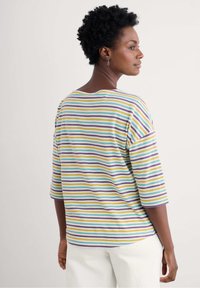 Camiseta de rayas de cuello redondo y mangas de tres cuartos. Los colores incluyen amarillo, verde, púrpura y azul sobre un fondo claro. Tejido suave.
